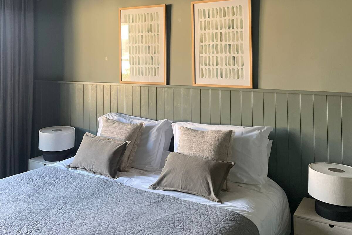 Quarto de hotel bem arrumado com uma cama, duas mesas de cabeceira, um abajur e duas fotografias emolduradas na parede.