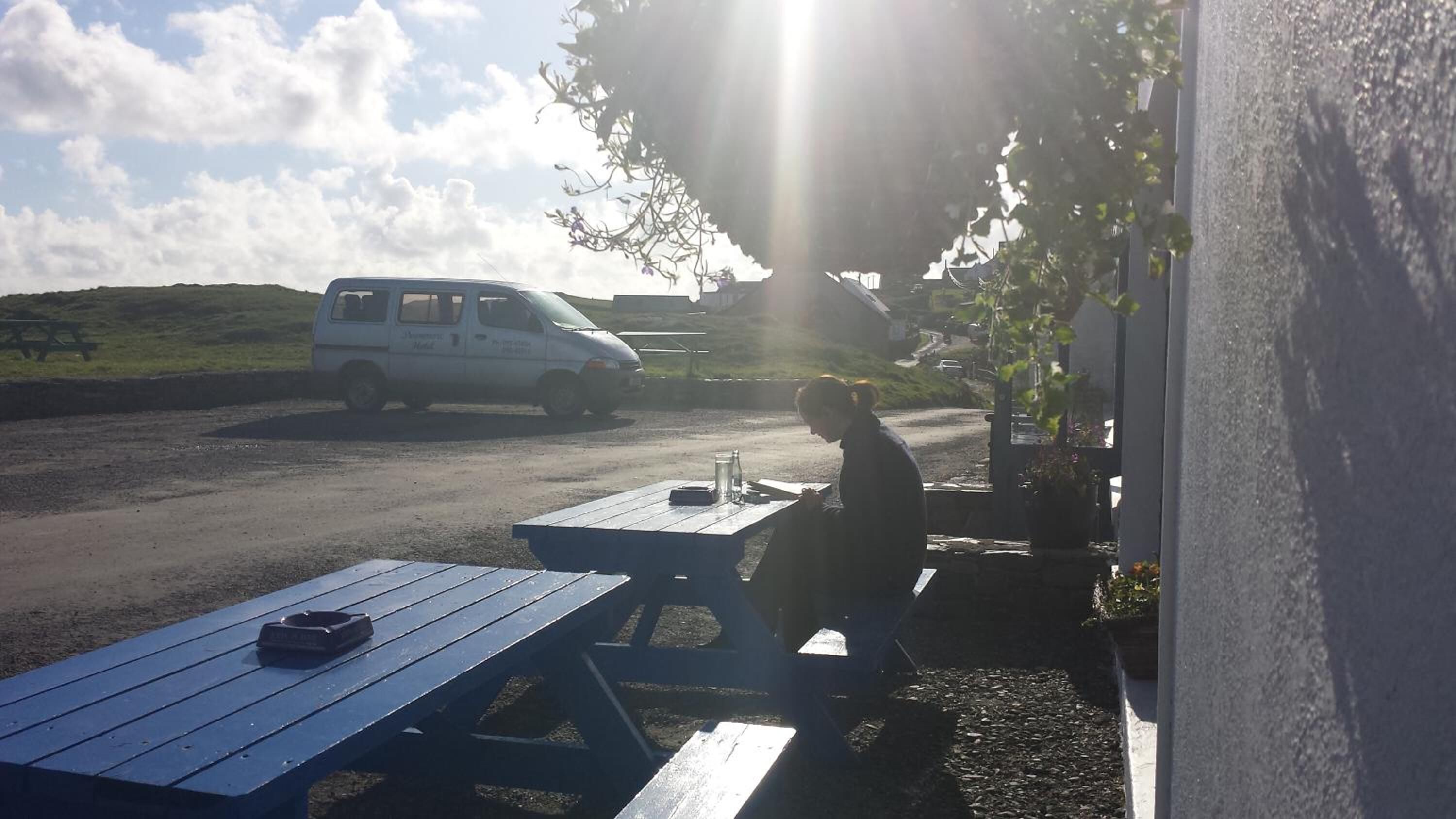 Una mesa de picnic con una botella y un recipiente, una camioneta blanca estacionada al borde de la carretera, y una persona sentada a la mesa.