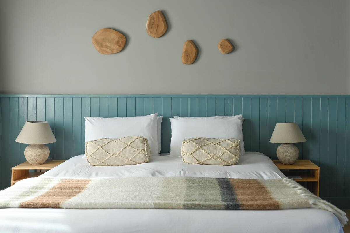 Uma cama com lençóis brancos, duas almofadas decorativas, um cobertor listrado, mesas de cabeceira de madeira com abajures e uma cabeceira azul-turquesa.