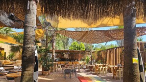 An outdoor seating area with wooden tables and benches, surrounded by palm trees and covered by a thatched roof. - No 22 Hotel Riders' Inn (Ula)