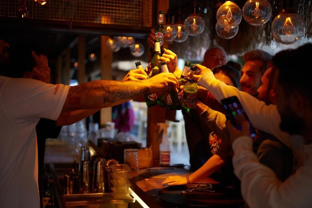 A bar setting with people toasting with bottles, a bartender, and a bar counter with various bottles and glasses.