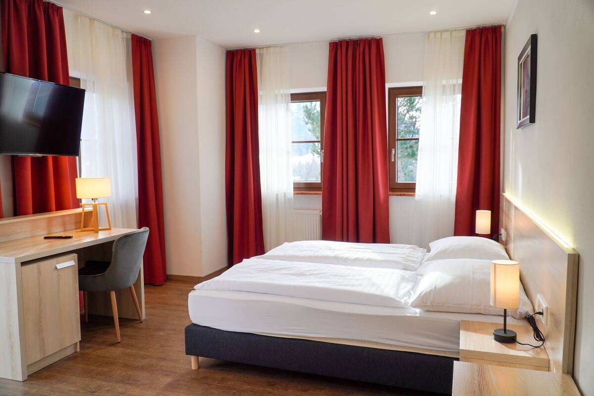 Habitación de hotel con cama, escritorio, silla, televisión y ventana con cortinas.
