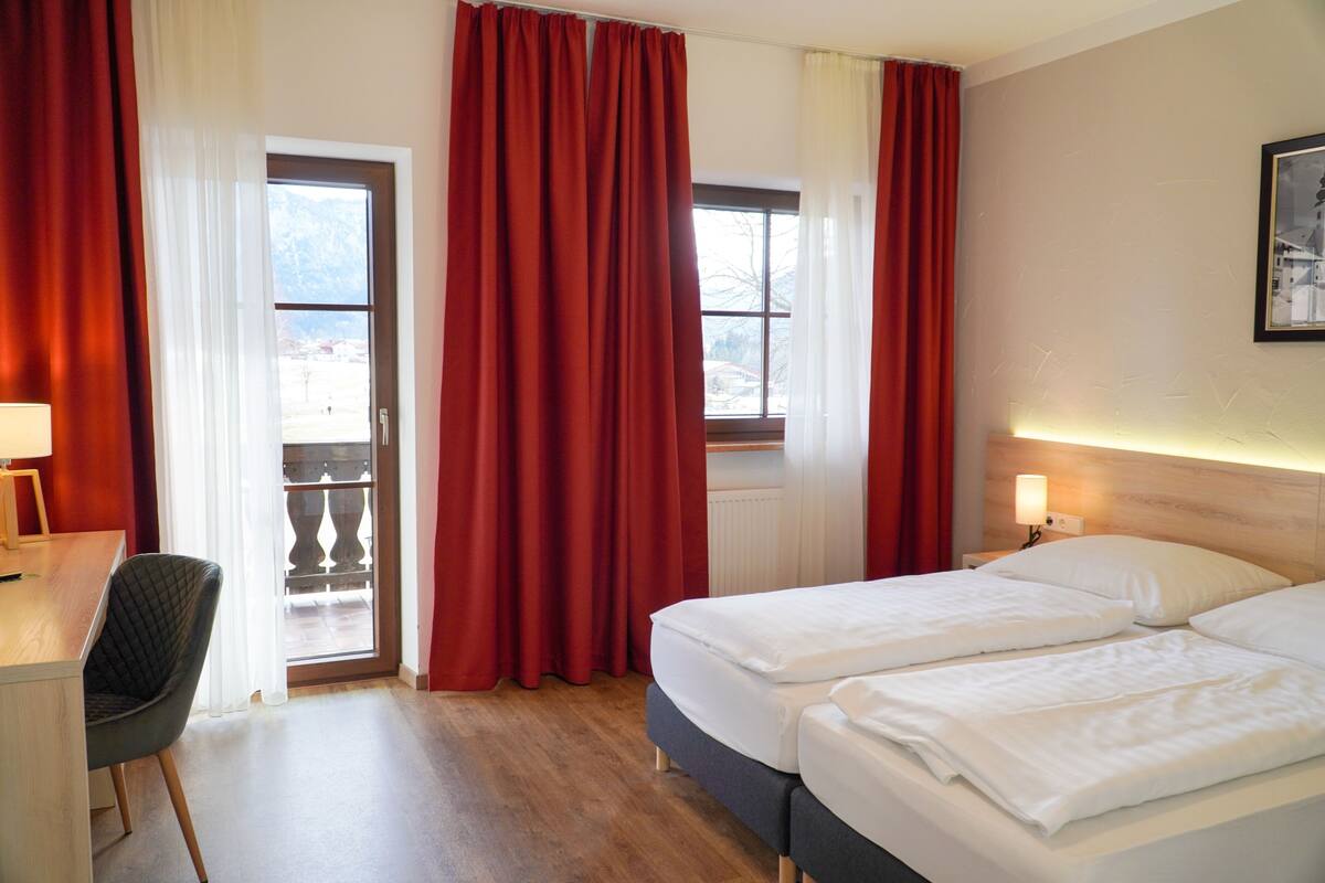Habitación de hotel con dos camas, un escritorio, una silla y un balcón con cortinas rojas.