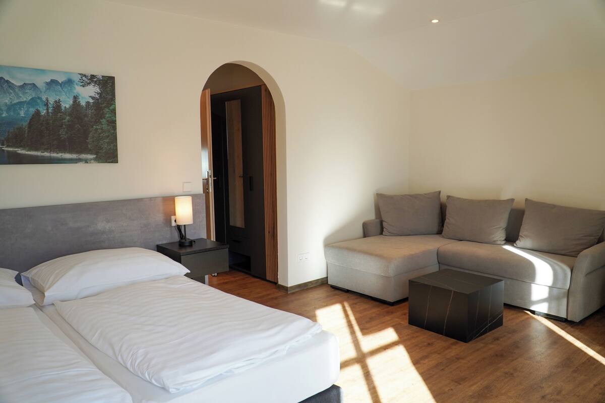 Una habitación de hotel moderna con una cama, un sofá, una mesita de noche y un cuadro de un paisaje montañoso.