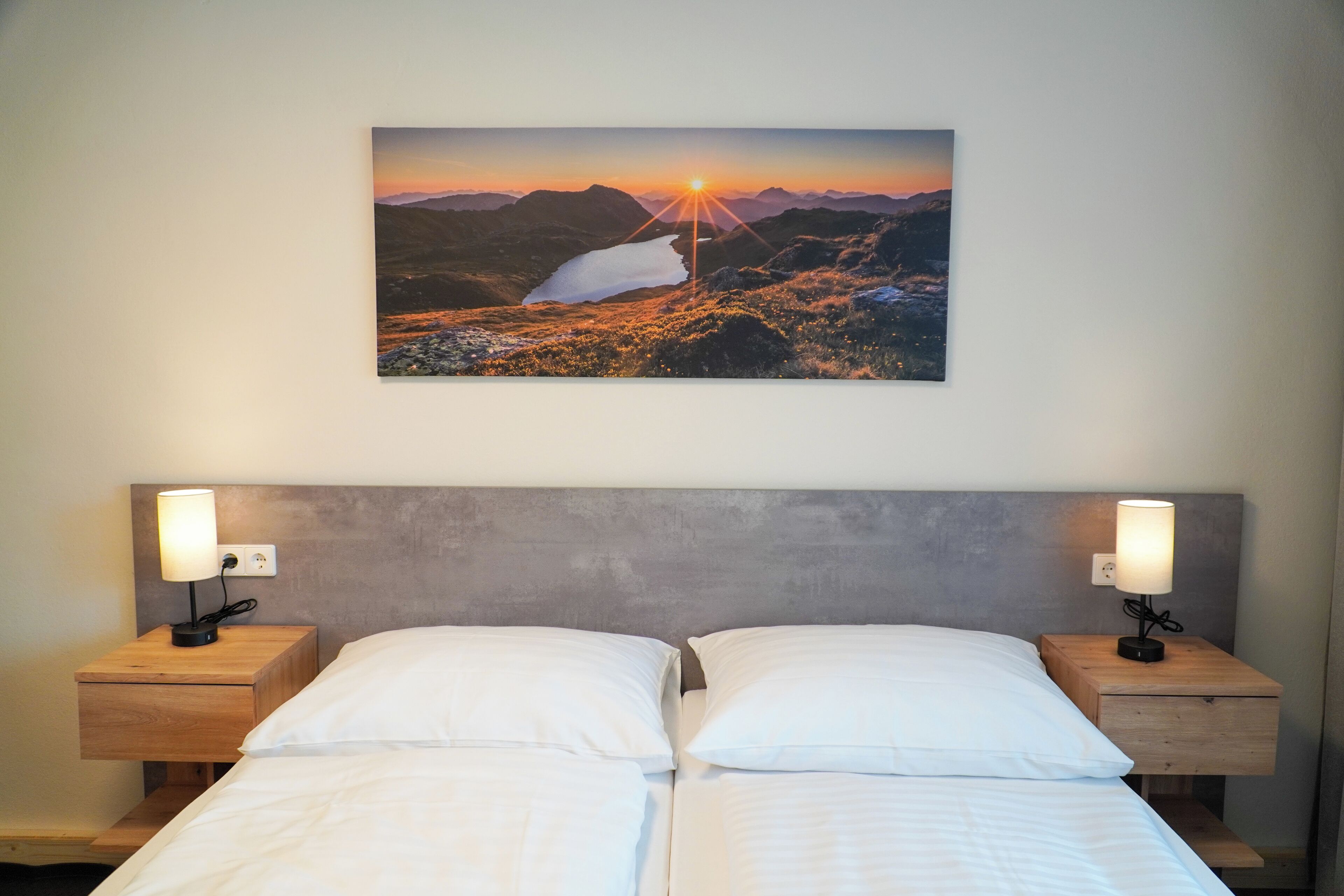 Un lit double avec du linge de lit blanc, deux tables de chevet avec des lampes et, au-dessus du lit, un tableau représentant un paysage au coucher du soleil.