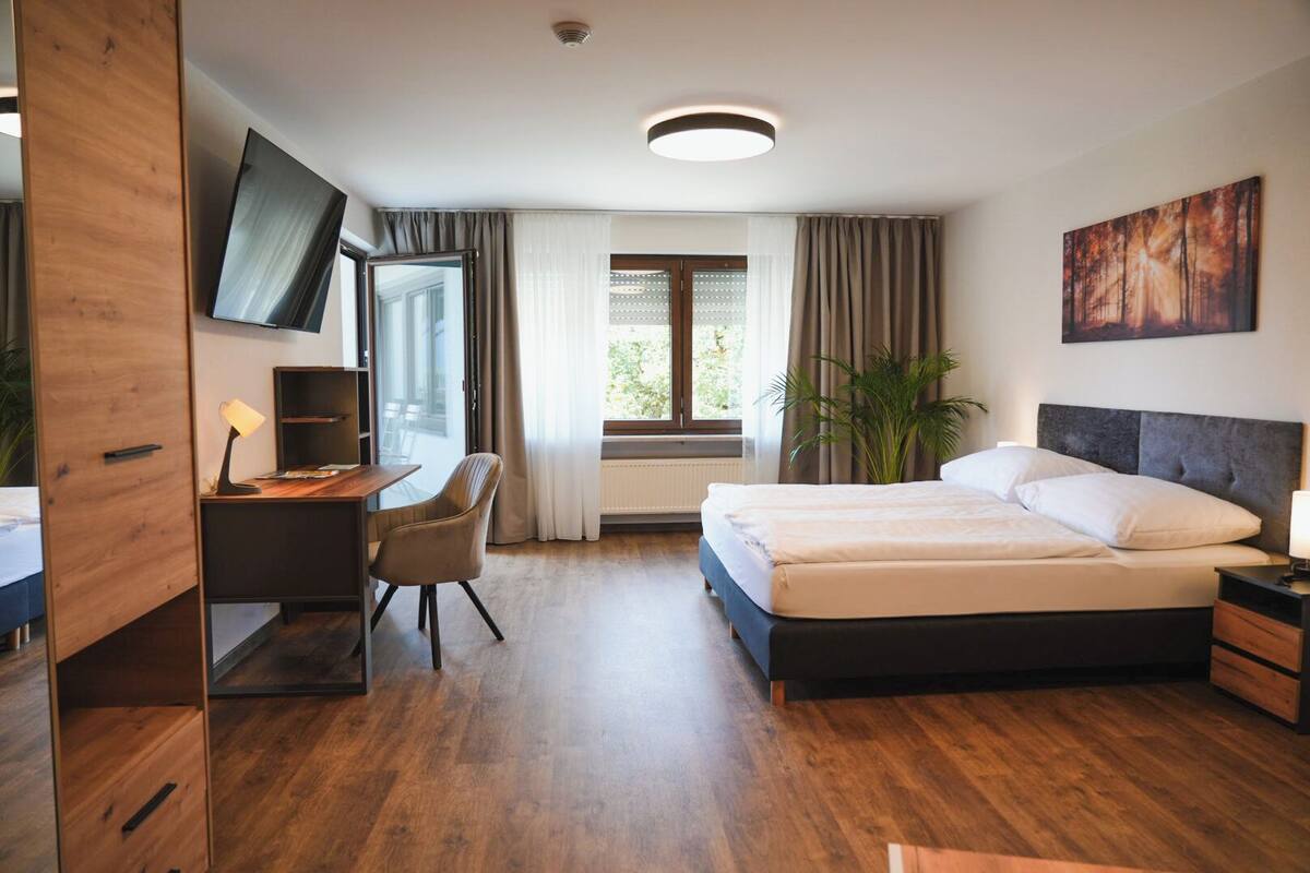 Una habitación de hotel moderna con un escritorio de madera, una cama con sábanas blancas, un televisor montado en la pared y un amplio ventanal con cortinas.