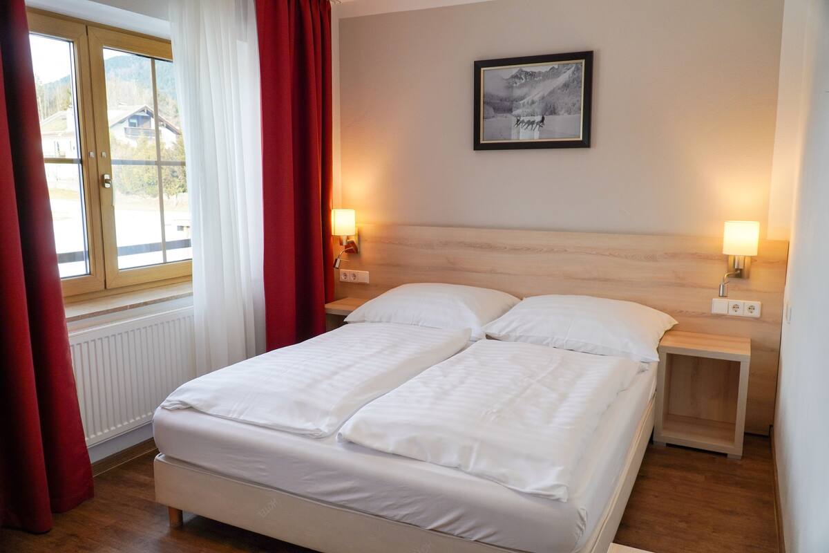 Cama doble con ropa de cama blanca, dos mesitas de noche con lámparas, un cuadro enmarcado en la pared y una ventana con cortinas rojas.