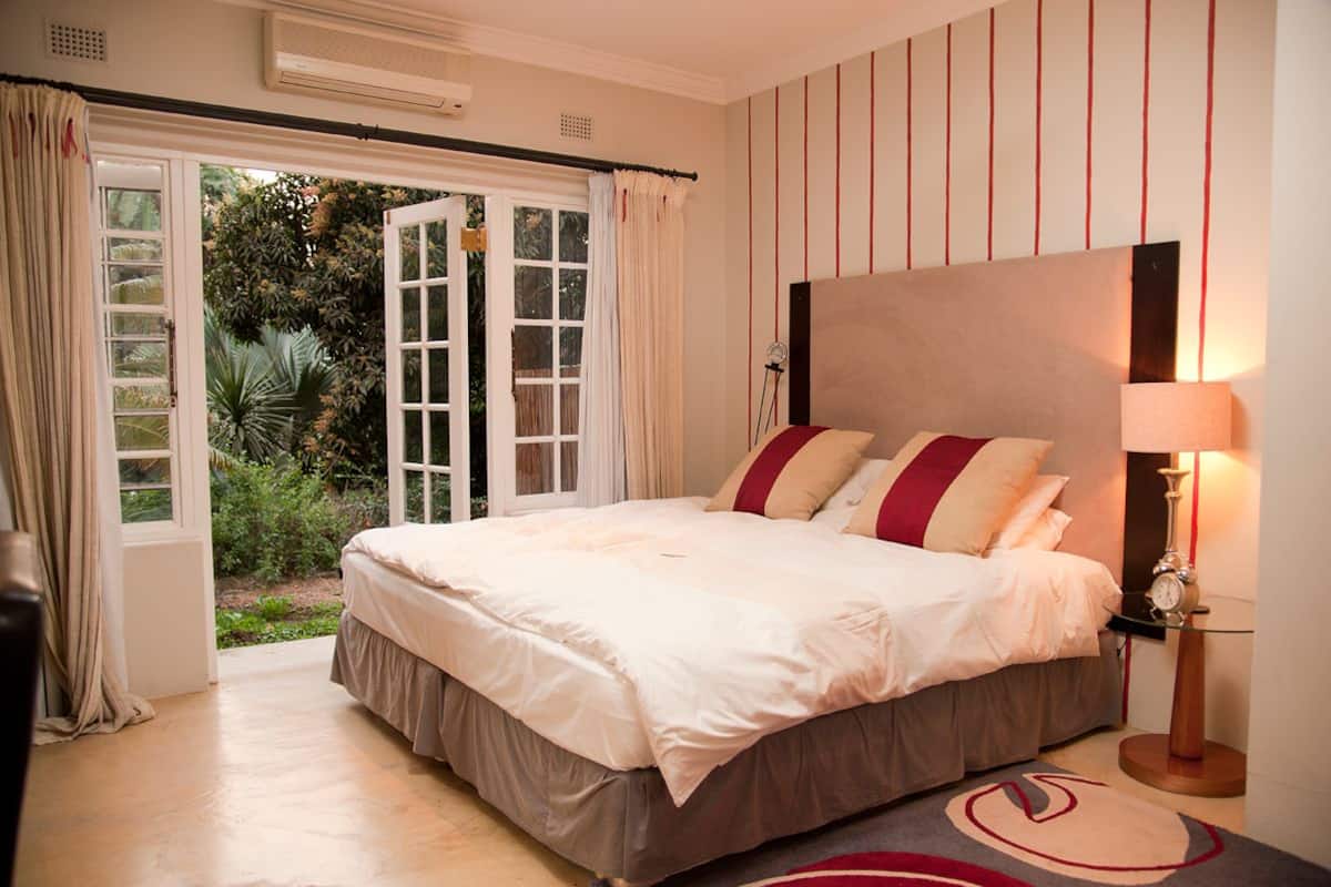 A bedroom with a large bed, a nightstand with a lamp, and a view of a garden through an open door.
