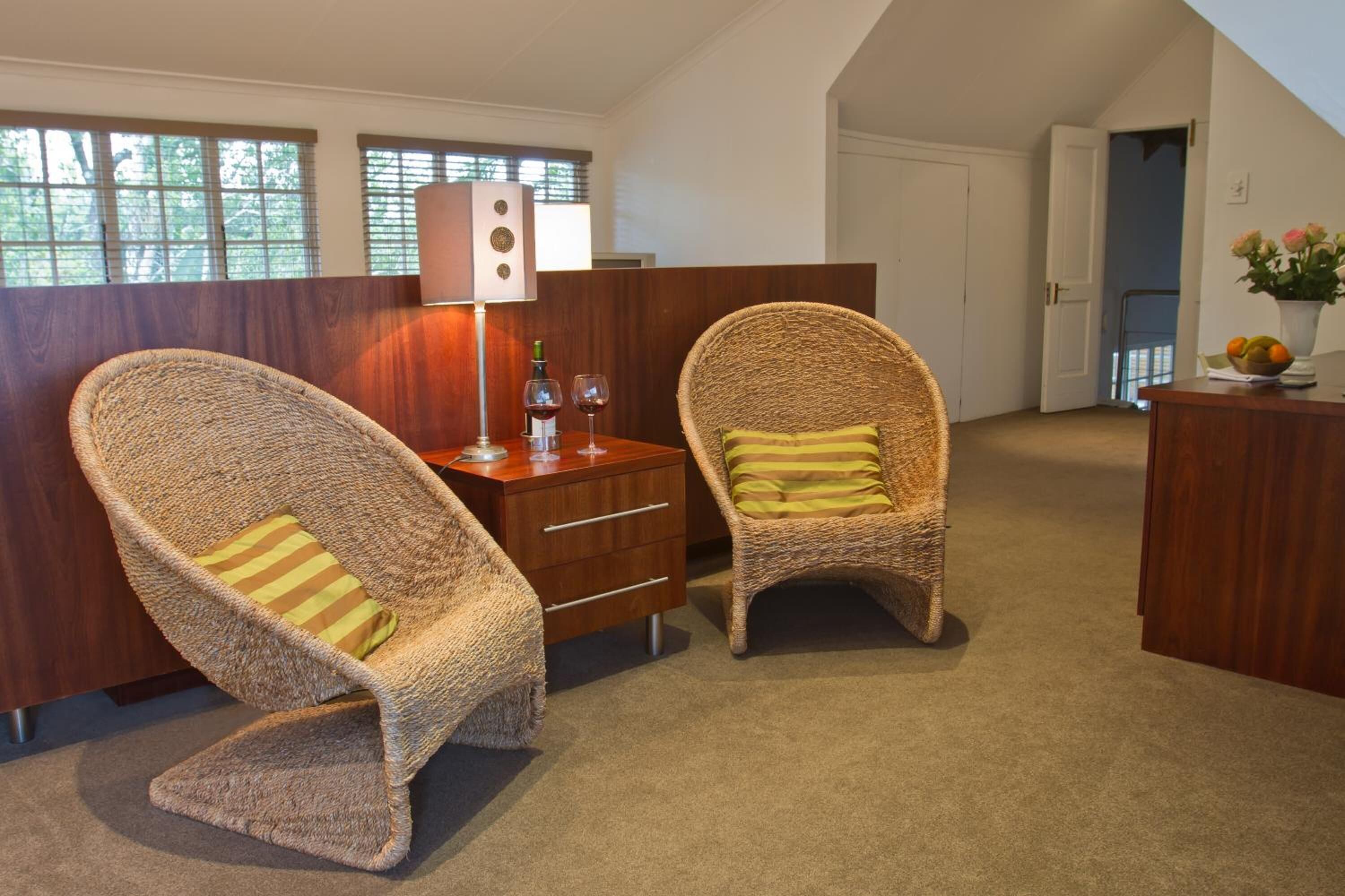 Quarto com duas cadeiras de vime, uma escrivaninha de madeira, um abajur e um vaso com flores.