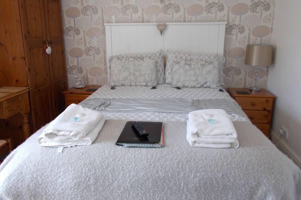 A neatly made bed with pillows and a blanket, a bedside table with a lamp, a wooden wardrobe, and a patterned wallpaper.