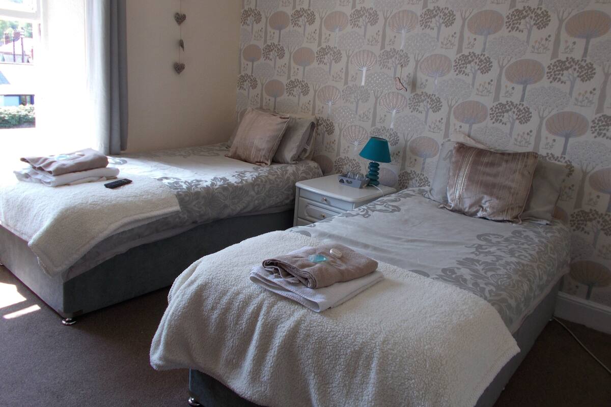 Two twin beds with towels, a nightstand with a lamp, and a wall with a tree pattern.
