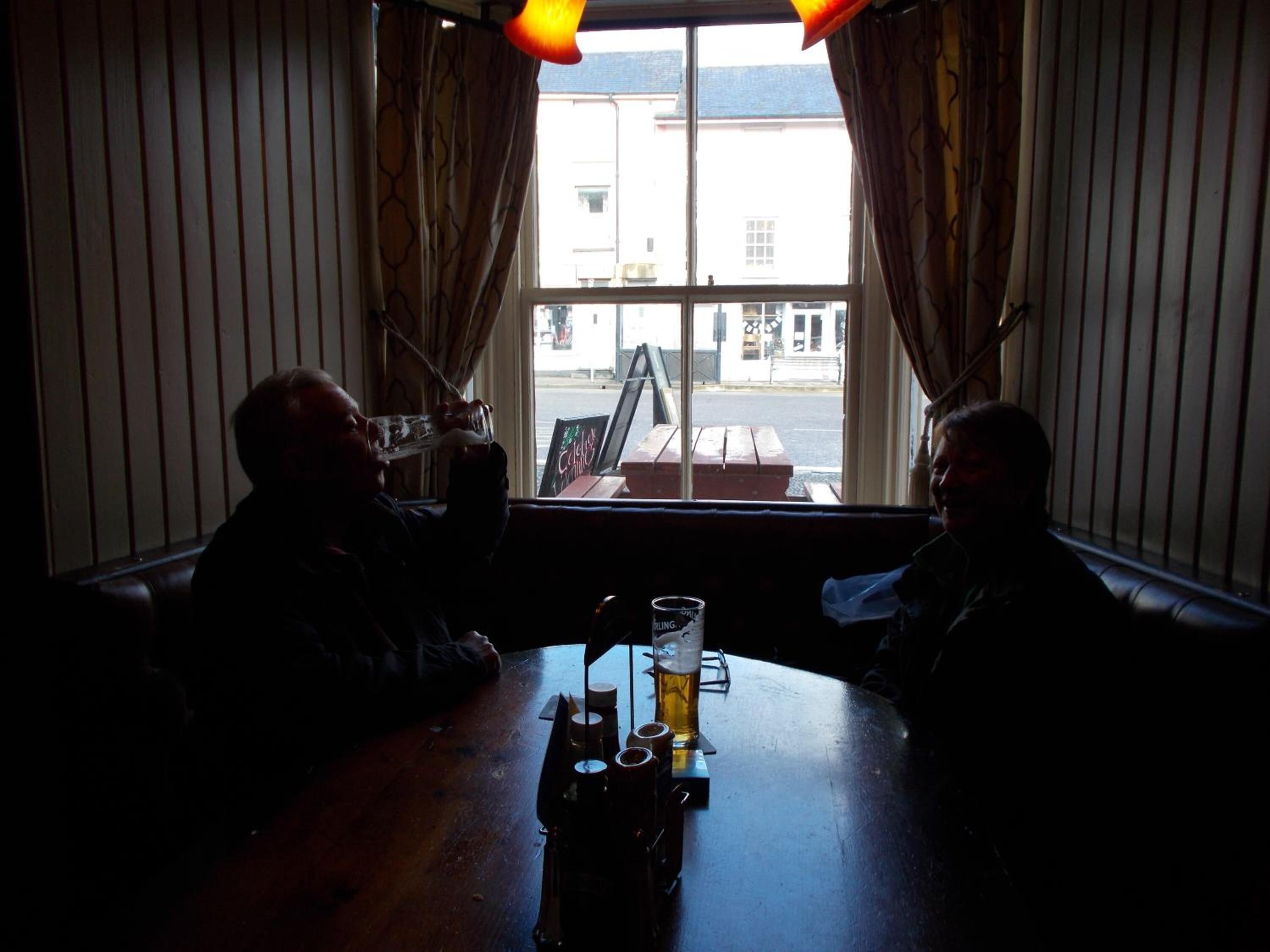 A dimly lit room with two people seated at a table, a window with curtains, and a view of the street outside.