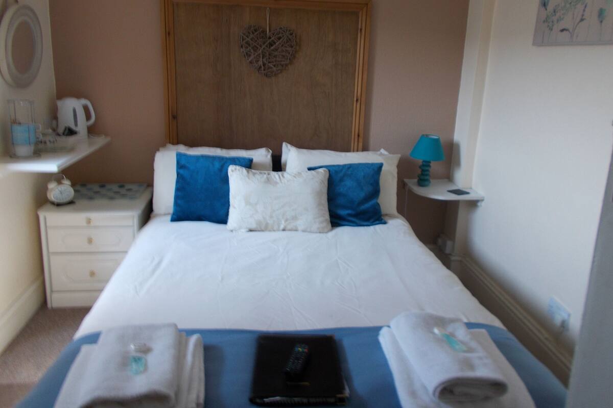 A neatly made bed with blue and white pillows, a heart-shaped decoration on the headboard, a bedside table with a lamp, and a small shelf with a kettle and cups.