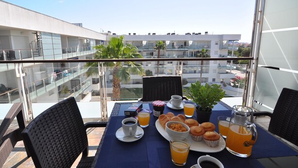 Een balkon met een ontbijttafel, compleet met muffins, koffie en sap, met uitzicht op een modern gebouwencomplex.