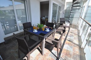 Un balcón con una mesa lista para el desayuno, sillas de mimbre y baranda de vidrio.