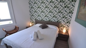 Ein ordentlich gemachtes Bett mit weißen Leinen und einem braunen Kopfkissen, ein Holzstuhl und eine Wand mit einem Blattmuster.