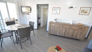 Ein modernes Wohnzimmer mit Essbereich, einem an der Wand montierten Fernseher, einer Holzkommode und einem kleinen Tisch mit Blumen.
