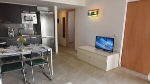 Ett modernt kök med mikrovågsugn, kylskåp och kaffemaskin. Det finns en matplats med ett bord för två personer, två stolar och en vas med blommor. En tv är monterad på ett skåp, och en trädörr syns.