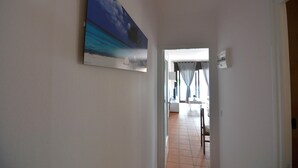 Een gang met een schilderij van een strand, een witte deur en een betegelde vloer die leidt naar een kamer met een bed en een raam.