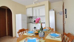 A dining area with a wooden table set for four, white cabinets in the background, and a kitchen area visible through an archway. - Vacation rental in Roses, Costa Brava: Cozy apartment 400 meters from the beach, capacity for 4 people, with terrace and amenities. (Rosas / Roses)