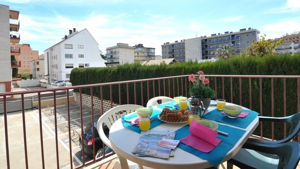 A balcony with a table set for breakfast, including croissants and orange juice, overlooking a residential area with buildings and a hedge. - Vacation rental in Roses, Costa Brava: Cozy apartment 400 meters from the beach, capacity for 4 people, with terrace and amenities. (Rosas / Roses)