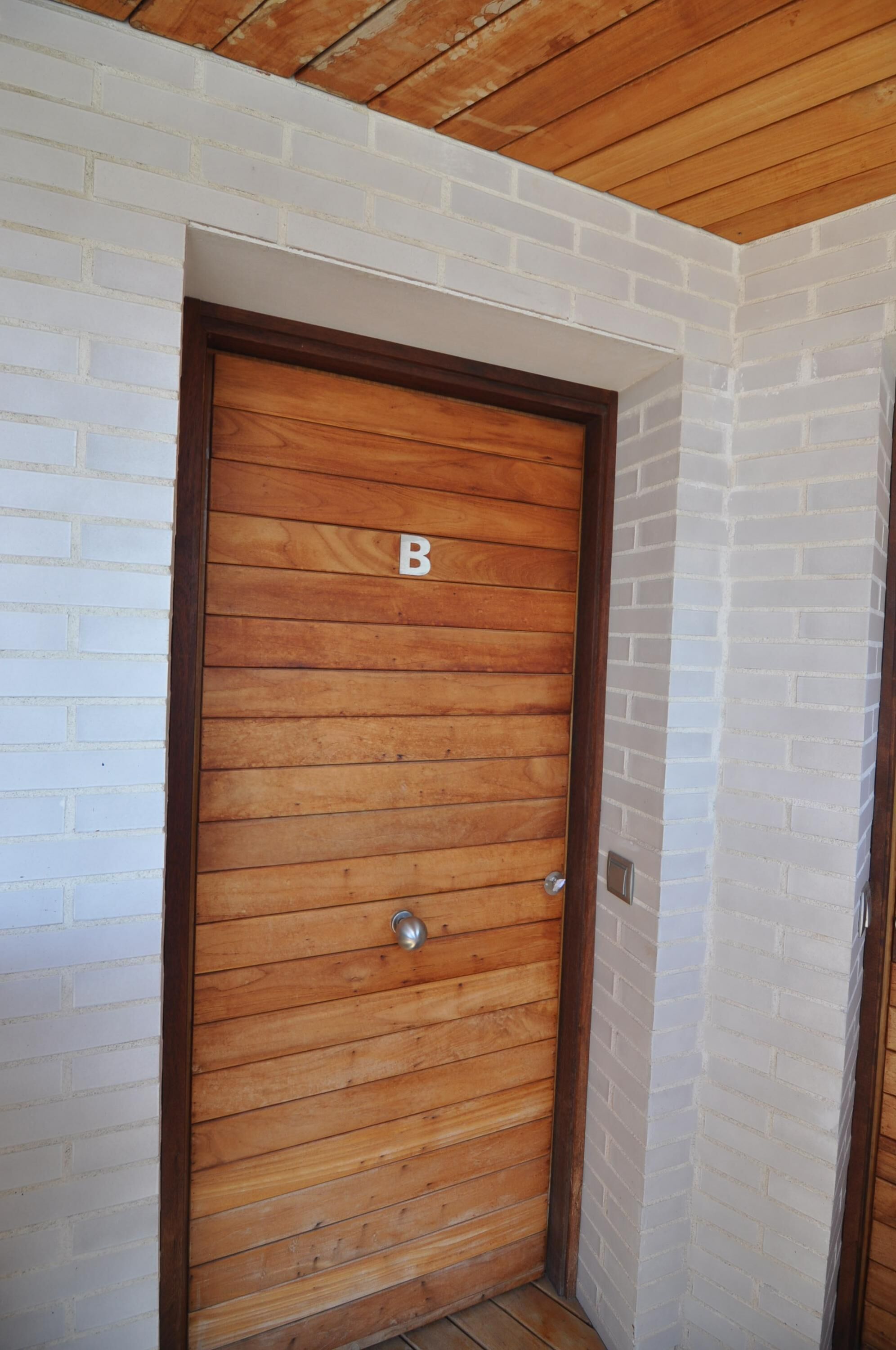 A wooden door with a letter 'B' on it, white brick walls, and a wooden ceiling.