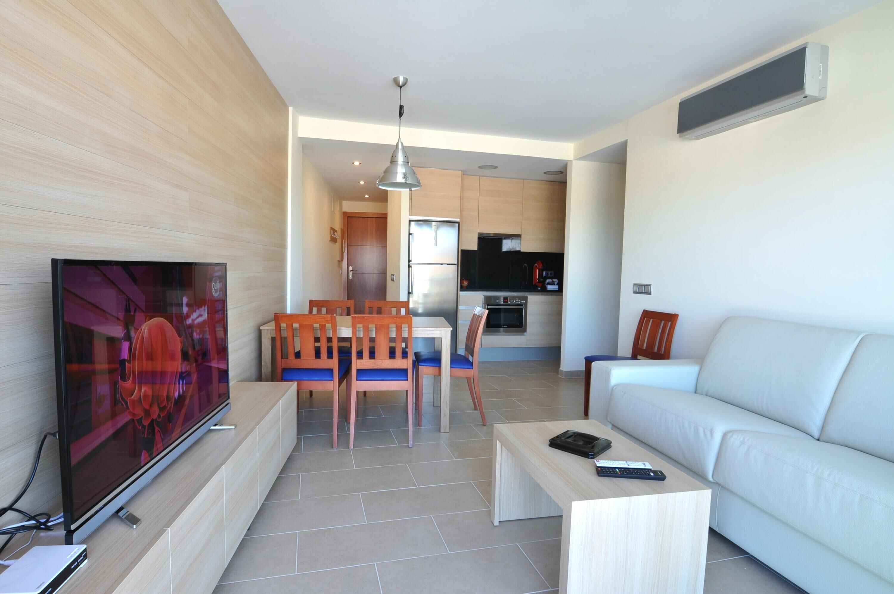 A modern living room with a flat-screen TV, wooden dining table, and chairs. There is a white sofa and a small coffee table. The kitchen is visible in the background with modern appliances.
