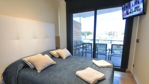A hotel room with a large bed, two pillows, a flat-screen TV mounted on the wall, and a balcony with chairs and a table.