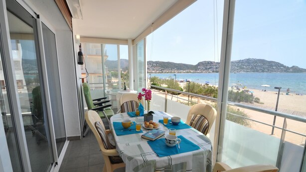 A balcony with a table set for two, overlooking a beach with a clear view of the sea and distant hills. - Charming apartment for rent in Roses, Costa Brava! Ideal for 5 people, just 10 meters from the beach. Book now! (Rosas / Roses)