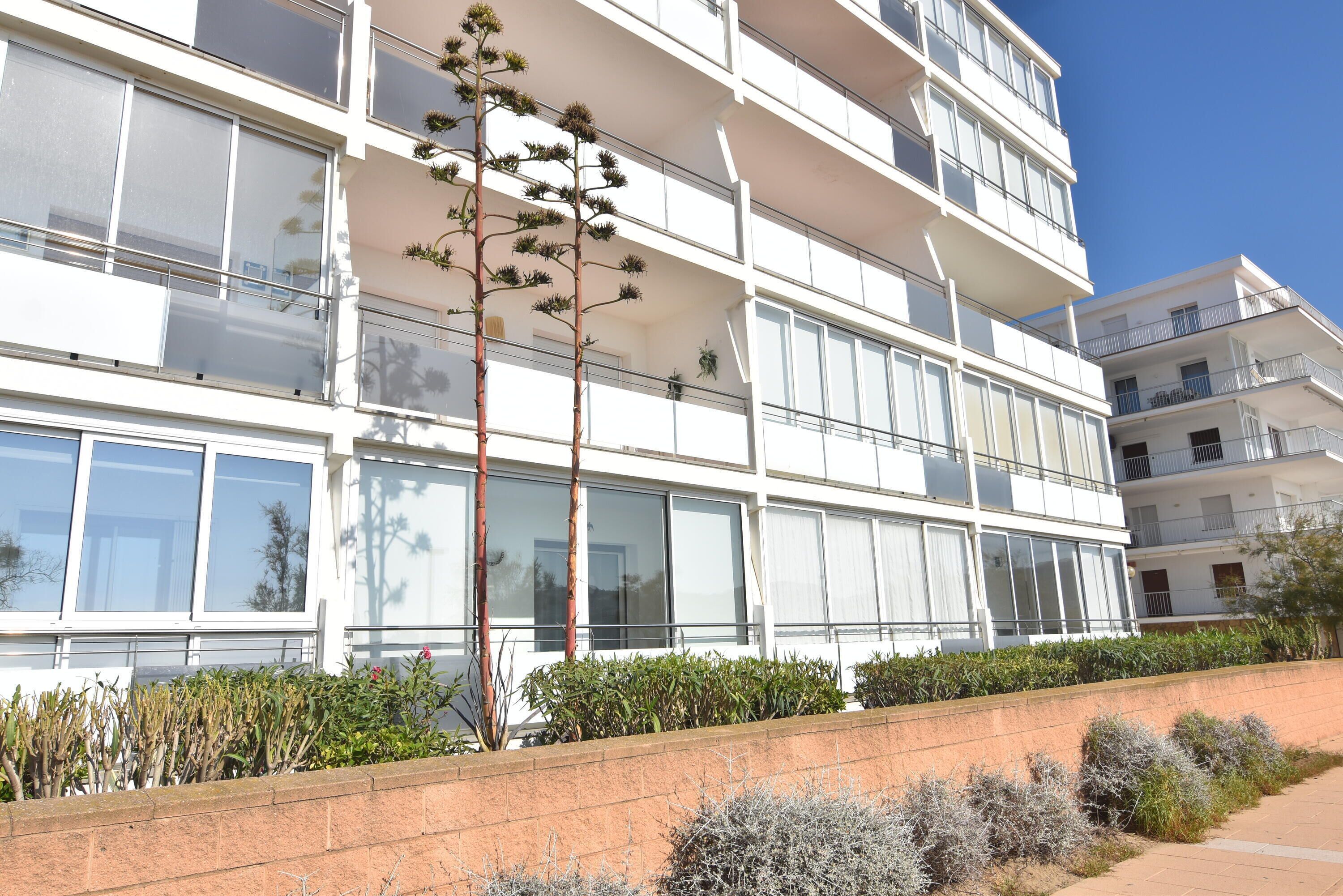 Um edifício residencial com vários andares, grandes janelas, um muro de suporte de tijolos e diversas plantas.