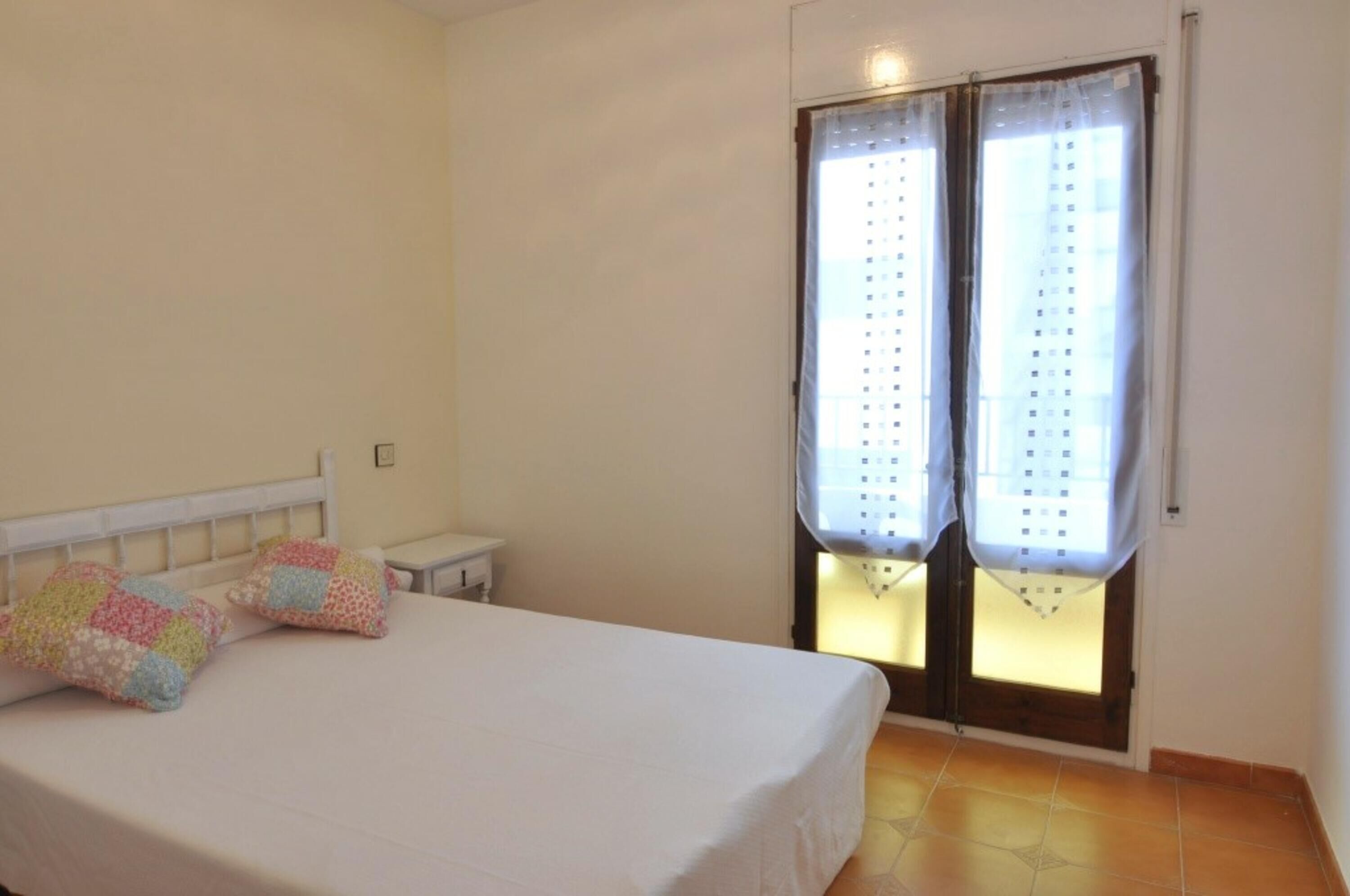 Un dormitorio con cama, dos almohadas, una mesita de noche y una ventana con cortinas.