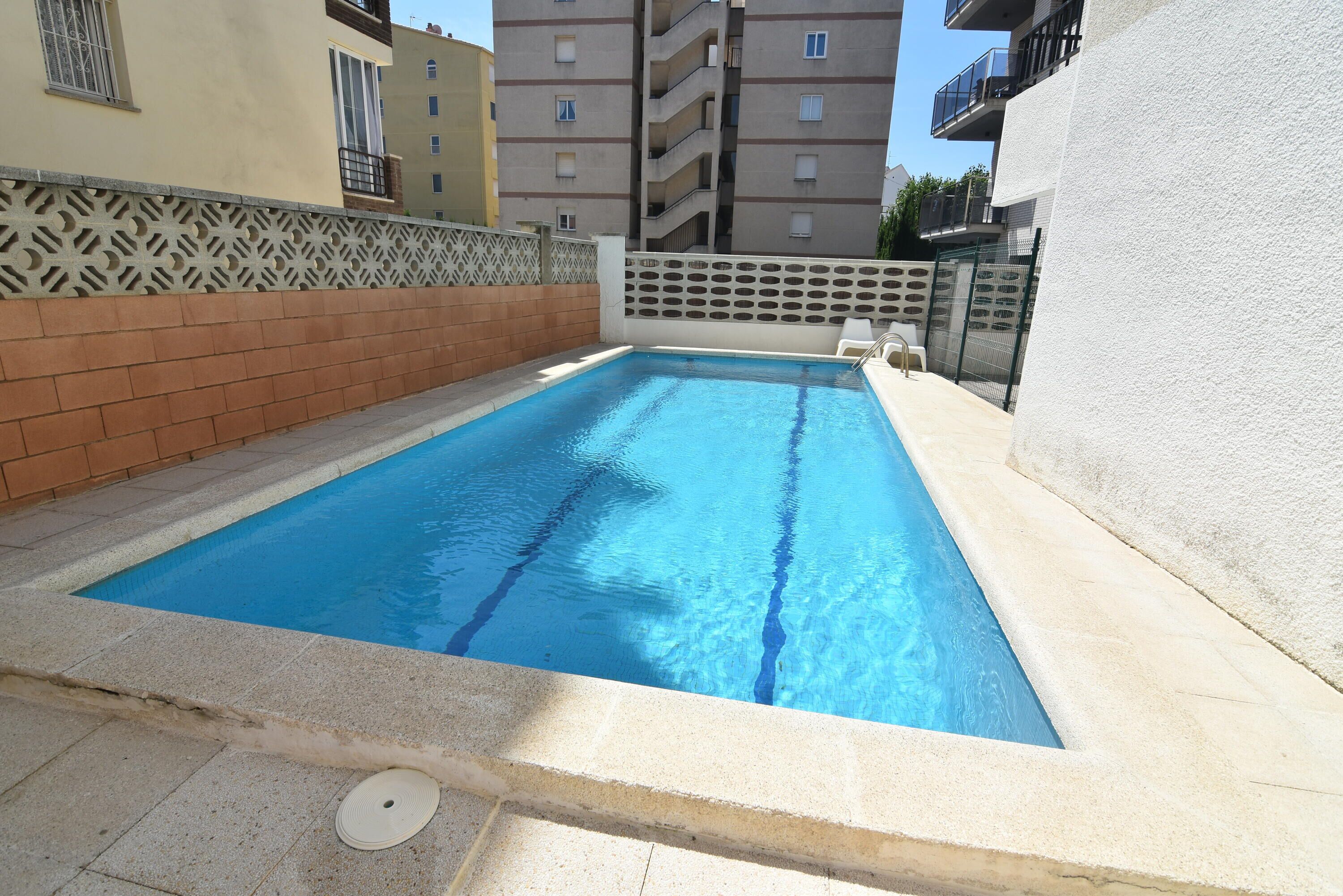 Una piscina rectangular con agua azul cristalina, rodeada por un solárium con baldosas y una cerca decorativa.