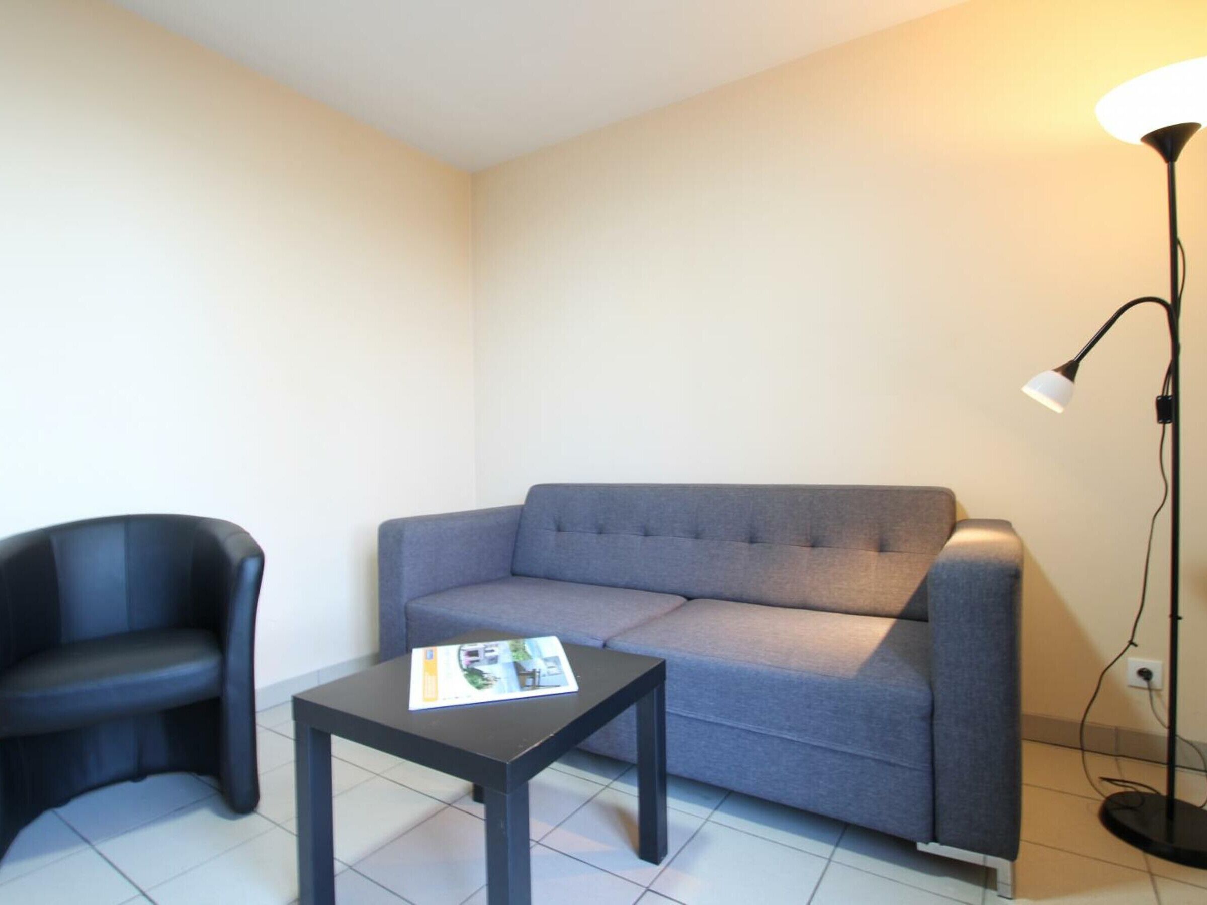 A small, modern room with a grey sofa, a black armchair, and a small table with a magazine. A floor lamp is visible in the corner.