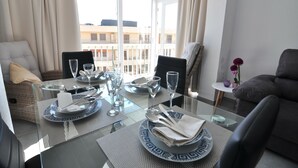 A dining area with a glass table set for two, a sofa, and a view of buildings through large windows.
