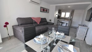 A modern living room with a grey sofa, a dining table set with plates and glasses, and a kitchen in the background.