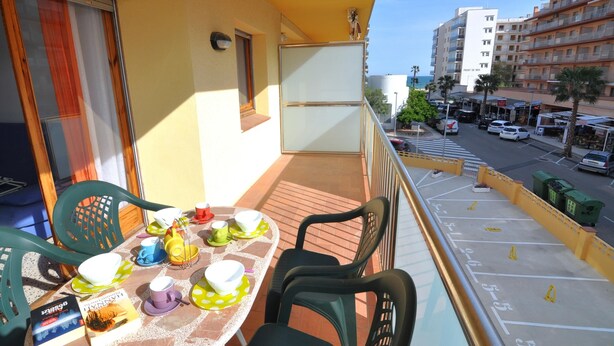 Uma varanda com mesa posta para refeições, cadeiras e janela com vista para a rua e prédios.