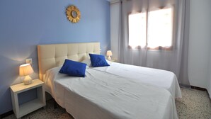 Cama bem arrumada com roupa de cama branca e duas almofadas azuis, uma mesa de cabeceira com abajur, um espelho fixado na parede e uma janela com cortinas.