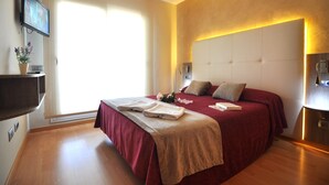 A hotel room with a large bed, a flat-screen TV mounted on the wall, a bedside table, and a window with blinds.