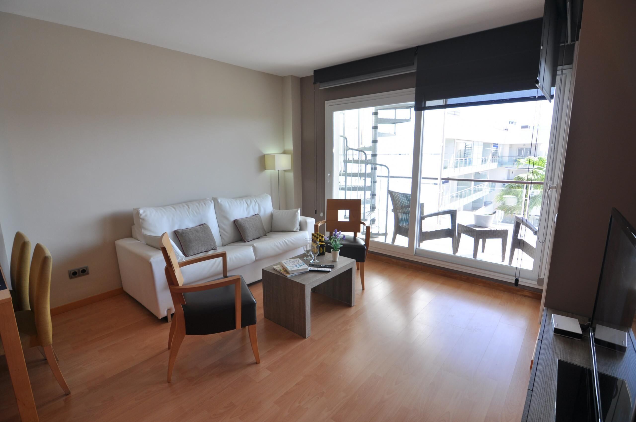 Un salón moderno con un sofá blanco, sillas de madera, una mesa de centro y un amplio ventanal corredero que da a un balcón con mobiliario exterior.