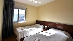 Ett hotellrum med två sängar, ett fönster med gardiner, ett nattduksbord och utsikt över staden.