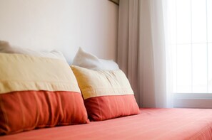 A bed with red bedding and two pillows, one red and one beige, against a white wall with a window and curtains. - Complejo Granaderos (Mendoza)