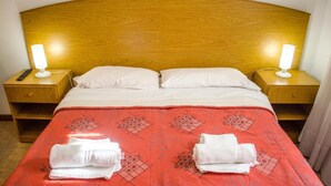 Ein Hotelzimmer mit hölzernem Kopfteil, zwei weißen Kissen, einer roten Decke mit geometrischem Muster, zwei weißen Handtüchern und Nachttischlampen.
