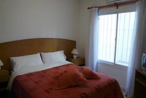 A single bed with a wooden headboard, white bedding, and a red blanket. There is a nightstand with a lamp to the left of the bed. A window with white curtains is visible to the right of the bed. - Complejo Granaderos (Mendoza)