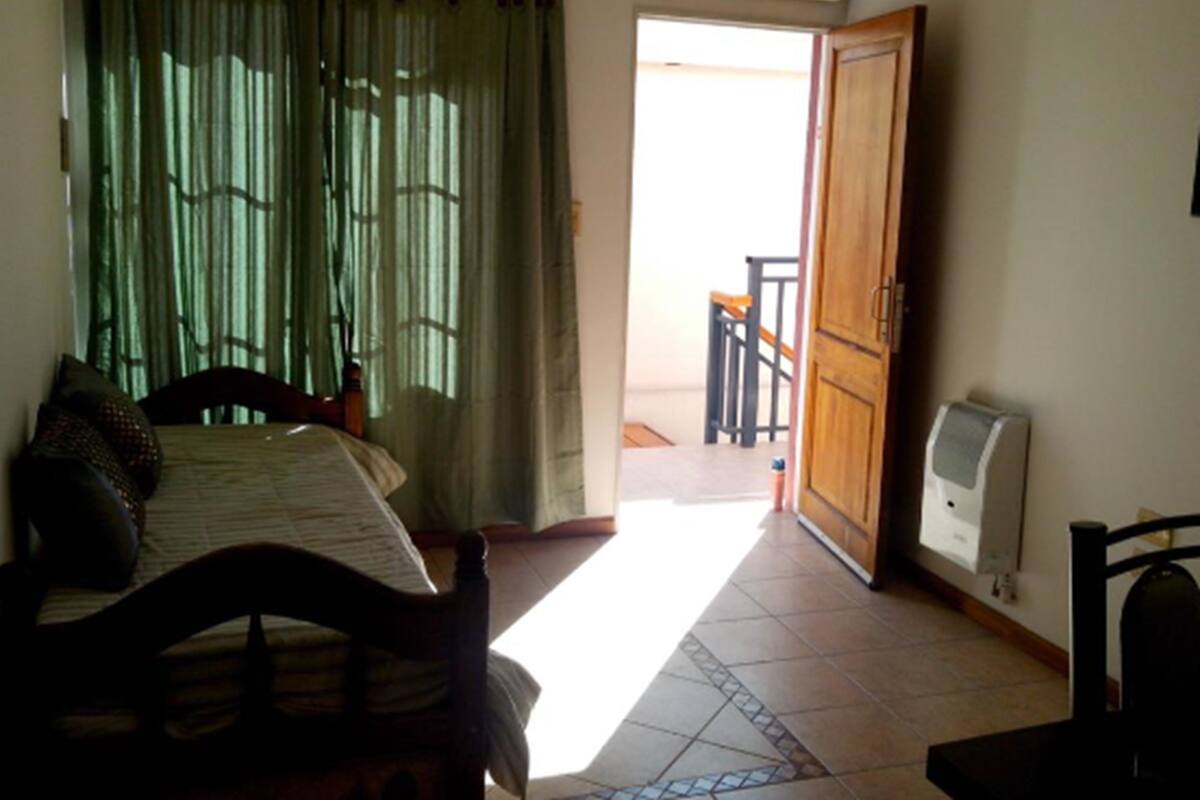 A bedroom with a bed, a wooden chair, a small table, a radiator, and a view of a balcony.