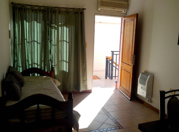 A bedroom with a bed, a wooden chair, a small table, a radiator, and a view of a balcony. - Complejo Granaderos (Mendoza)