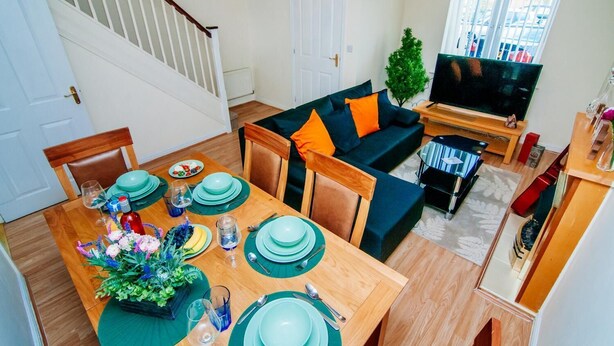 A dining area with a wooden table set for four, a sofa with orange cushions, a TV, and a staircase. - Harris House (Doncaster)