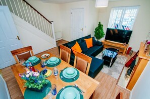 A dining area with a wooden table set for four, a sofa with orange cushions, a TV, and a staircase. - Harris House (Doncaster)