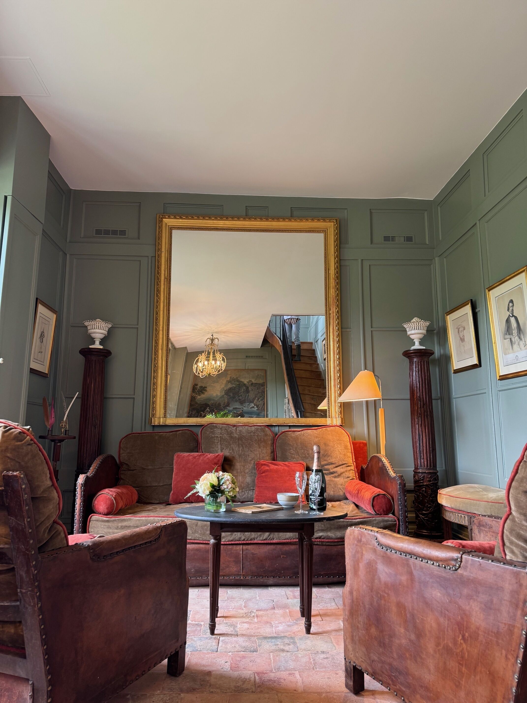 A room with a large mirror, a sofa, armchairs, a small table with a vase and drink, and a lamp.