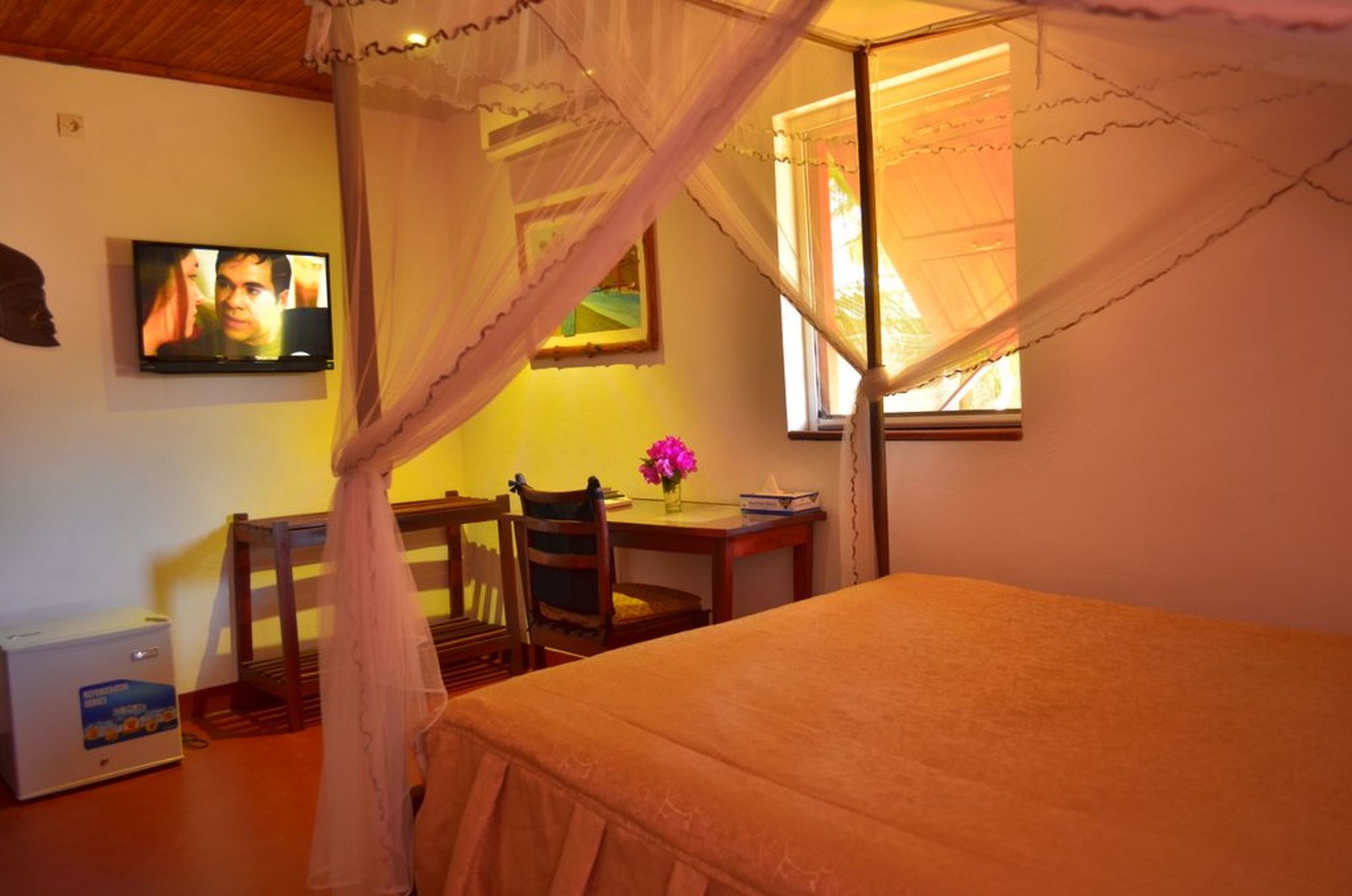 A bedroom with a canopy bed, a television on the wall, a desk with a chair, and a window with curtains.