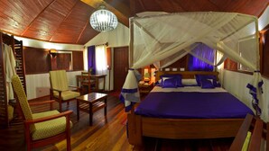 A room with a wooden ceiling, a bed with a canopy, a chair, a table, and a lamp. - Edena Kely (Mahajanga)
